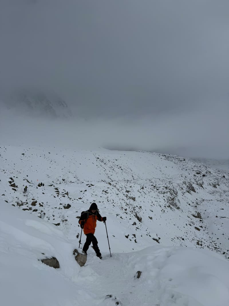 A trekker in an orange jacket descending a snowy slope under heavy cloud