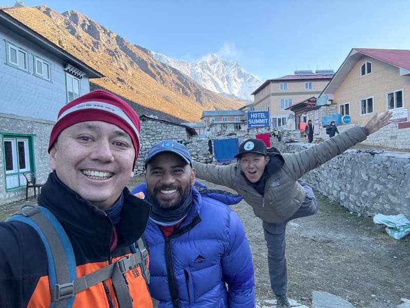 Selfie of David and Praveen in down jackets with Furi Sherpa balancing on one leg, arms outstretched, among the lodges of Dingboche with a snowy peak behind