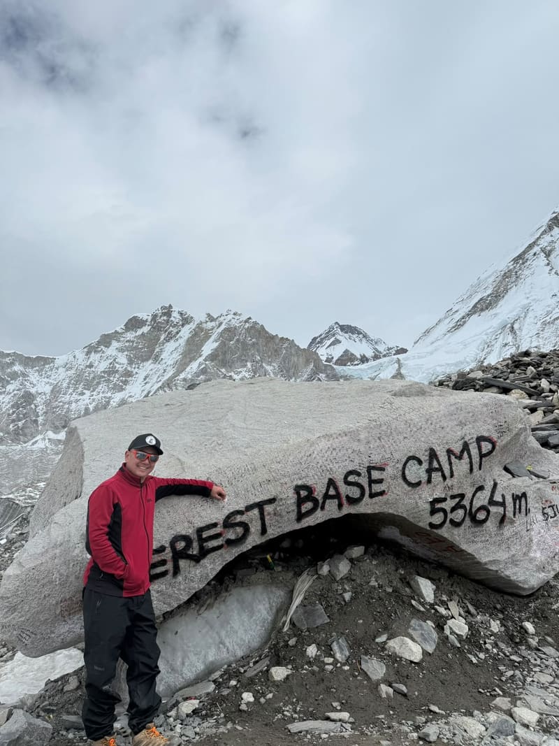 David in a red jacket leaning on the Everest Base Camp rock, painted 5364 m, with snowy peaks under cloud