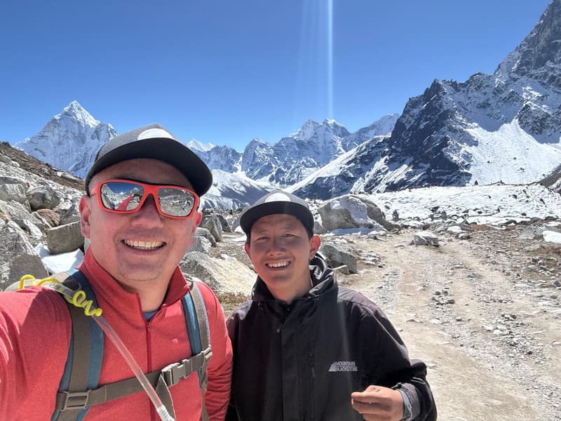 Selfie of David in a red top and orange sunglasses with Furi Sherpa on a rocky trail, both in matching caps, Ama Dablam and snowy peaks behind under a clear blue sky