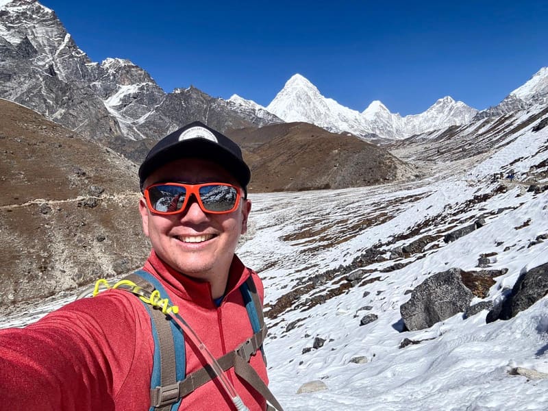 Selfie of David in a red top and cap on a snowy slope, a white pyramid peak at the head of the valley under a deep blue sky