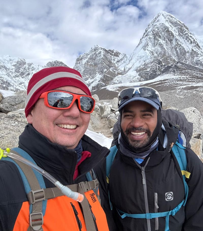 Selfie of David in a red and grey beanie with Praveen, both smiling, in front of a steep snow-covered peak